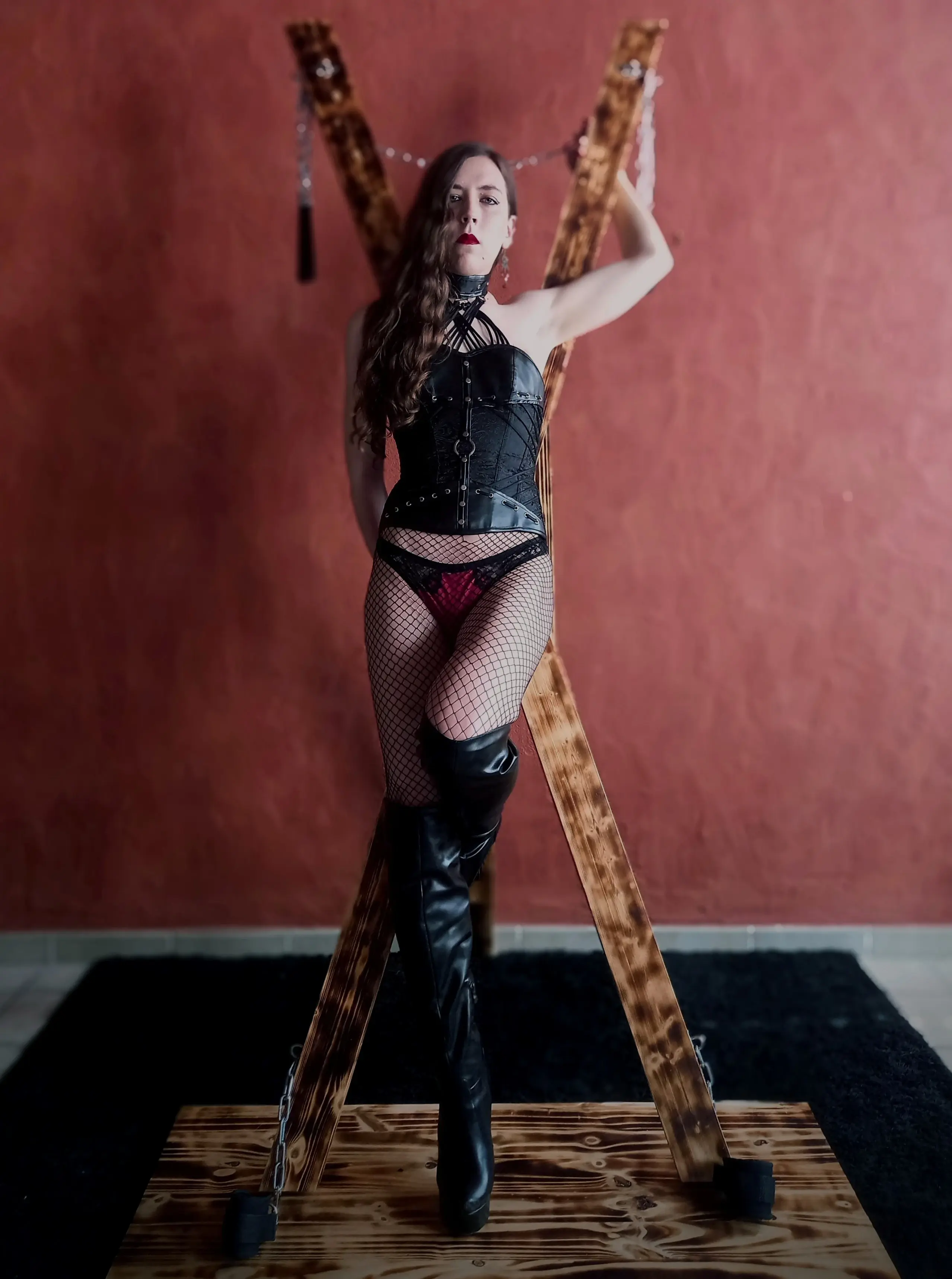 Gabrielle, le regard sévère, portant un corset et des cuissardes effet cuir, devant sa croix de Saint André